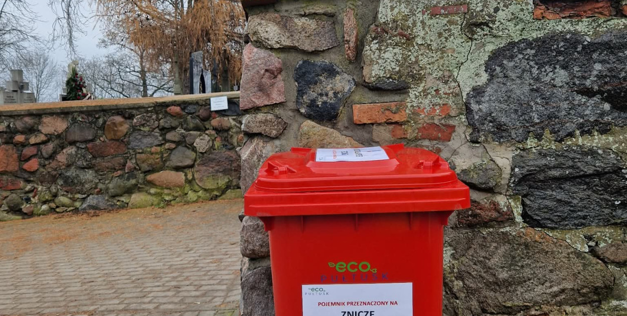 Czerwony pojemnik na zużyte znicze elektryczne ustawiony przy wejściu na cmentarz w Pułtusku.